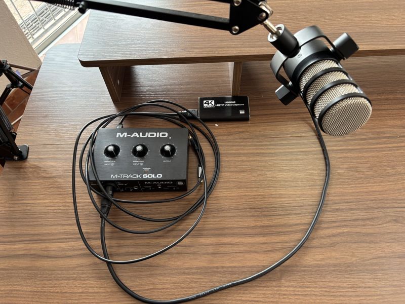 Rode PodMic on a boom arm next to an M-Audio M-Track Solo interface and a USB HD video capture device on the desk