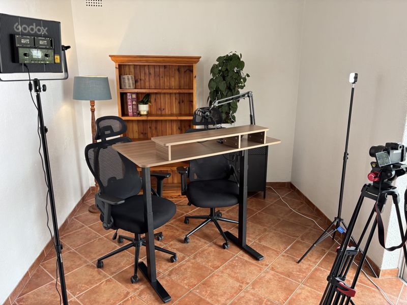 Desk raised to standing height with microphone boom arm and two chairs in front of a pine bookshelf, with a Godox LED panel and a small LED light on stands either side