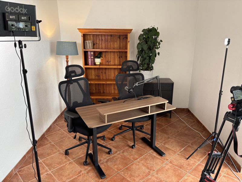 The studio room: a desk at seated height with a Rode PodMic on a boom arm, two office chairs, a pine bookshelf, a floor lamp and a plant, with a Godox panel light on the left and a camera on a tripod on the right